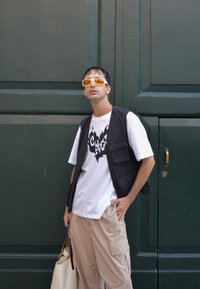 Chaleco ligero negro sobre una camiseta blanca con un gráfico negro, pantalones cargo beige y gafas de sol amarillas, sosteniendo una bolsa de tela color crema. Fondo verde.