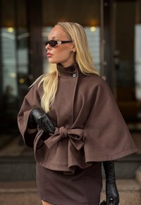 Ξανθιά γυναίκα με καφέ παλτό κάπα με φιόγκο, μαύρα δερμάτινα γάντια και μαύρα γυαλιά ηλίου, που στέκεται μπροστά σε μια θολή είσοδο κτιρίου.