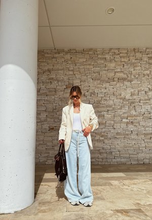 Blazer crema sobre una camiseta blanca, jeans de pierna ancha en un tono azul claro y zapatillas blancas. Sostenido un bolso marrón oscuro; de pie contra una pared de piedra.