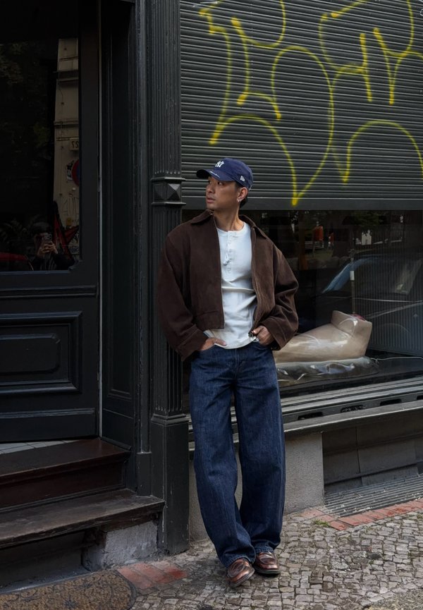 Uomo con berretto blu, giacca marrone, camicia bianca e jeans appoggiato a un edificio con una serranda metallica coperta di graffiti.