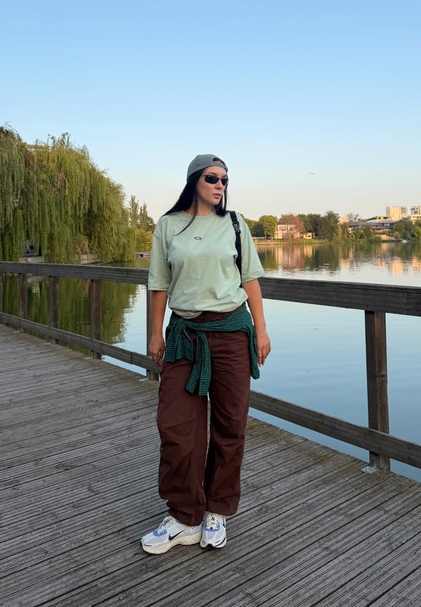 Giovane donna con occhiali da sole e abbigliamento casual in piedi su un molo di legno vicino a un lago tranquillo, con alberi e edifici sullo sfondo.