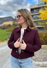 Burgundi színű, gallérral ellátott, rövidített dzseki, elülső zsebekkel és patentos záródással, világosszürke felsővel és magas derekú kék farmernadrággal párosítva.