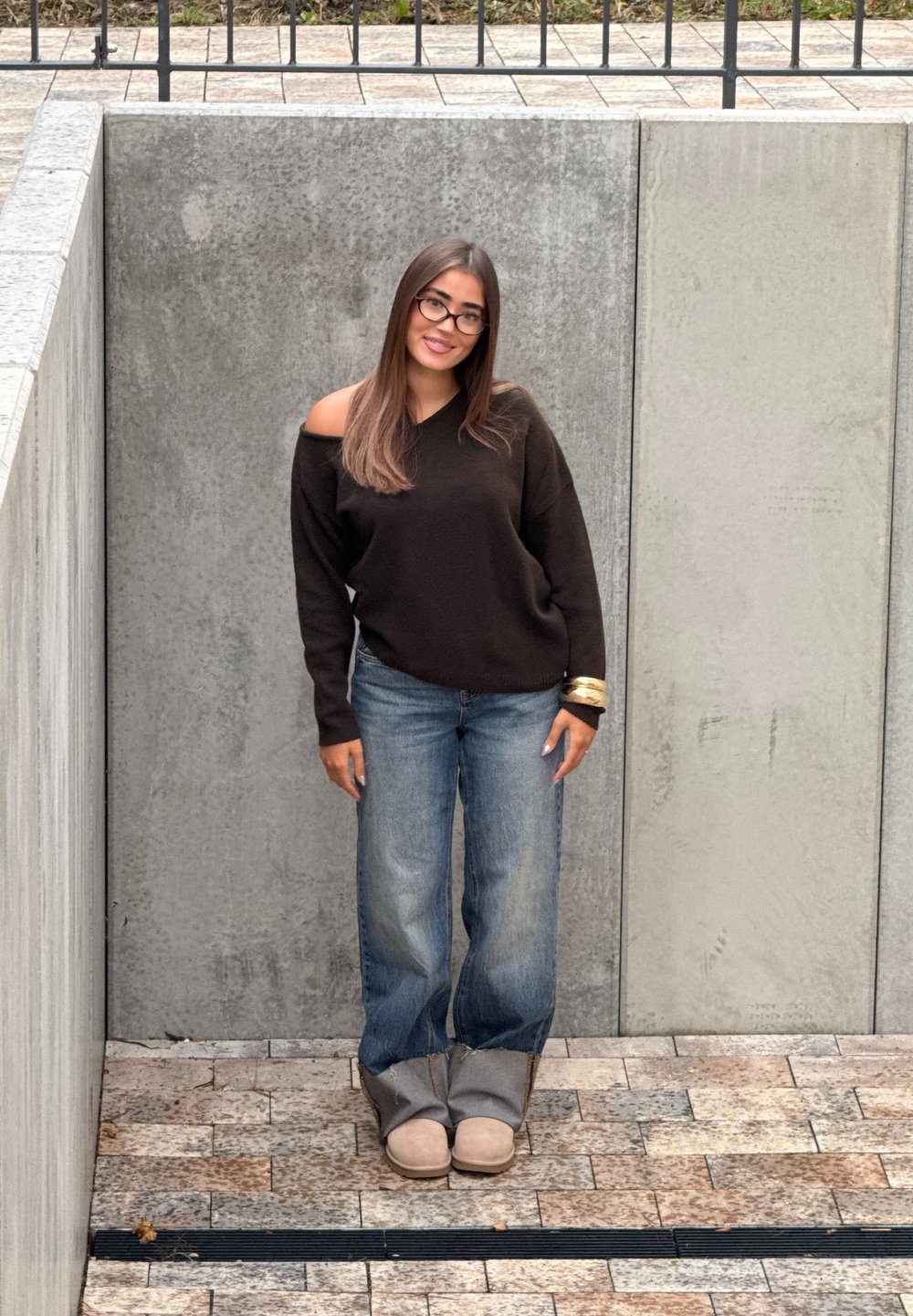 Jonge vrouw met lang haar en een bril staat glimlachend, gekleed in een off-shoulder zwarte trui, blauwe jeans en beige laarzen tegen betonnen muren.