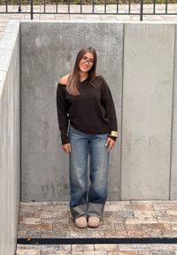 Jonge vrouw met lang haar en een bril staat glimlachend, gekleed in een off-shoulder zwarte trui, blauwe jeans en beige laarzen tegen betonnen muren.