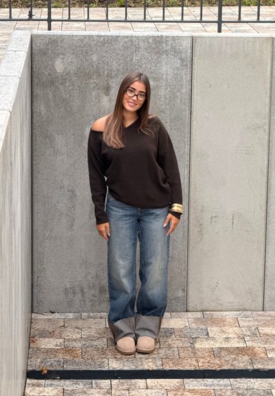 Joven con cabello largo y gafas sonríe, vistiendo un suéter negro de hombros descubiertos, jeans azules y botas beige, frente a paredes de concreto.
