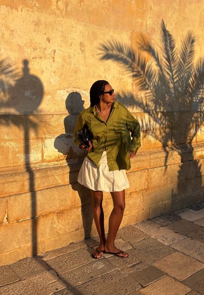 Camisa de satén verde oliva, falda blanca con volantes, bolso negro y chanclas frente a una pared de piedra texturizada con sombras de palmeras.