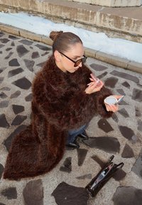 Femme en manteau de fourrure marron appliquant du rouge à lèvres tout en tenant un miroir compact, assise sur un sol pavé de pierres à côté d'un sac à main ouvert.