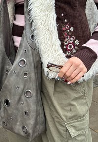 Une main tenant des lunettes écaille de tortue, portant un pull rayé, un gilet brodé de fleurs avec bordure en fourrure blanche, un pantalon cargo vert et un sac gris avec des anneaux métalliques.