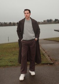 Jeune homme en pull gris, manteau foncé et pantalon marron marchant près d'un lac par une journée nuageuse, les mains dans les poches.