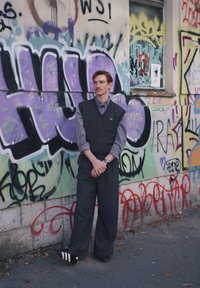 Noor mees halli särgi, tume vesti ja laiade pükstega toetub linnatänaval grafitiga kaetud müüri äärde.