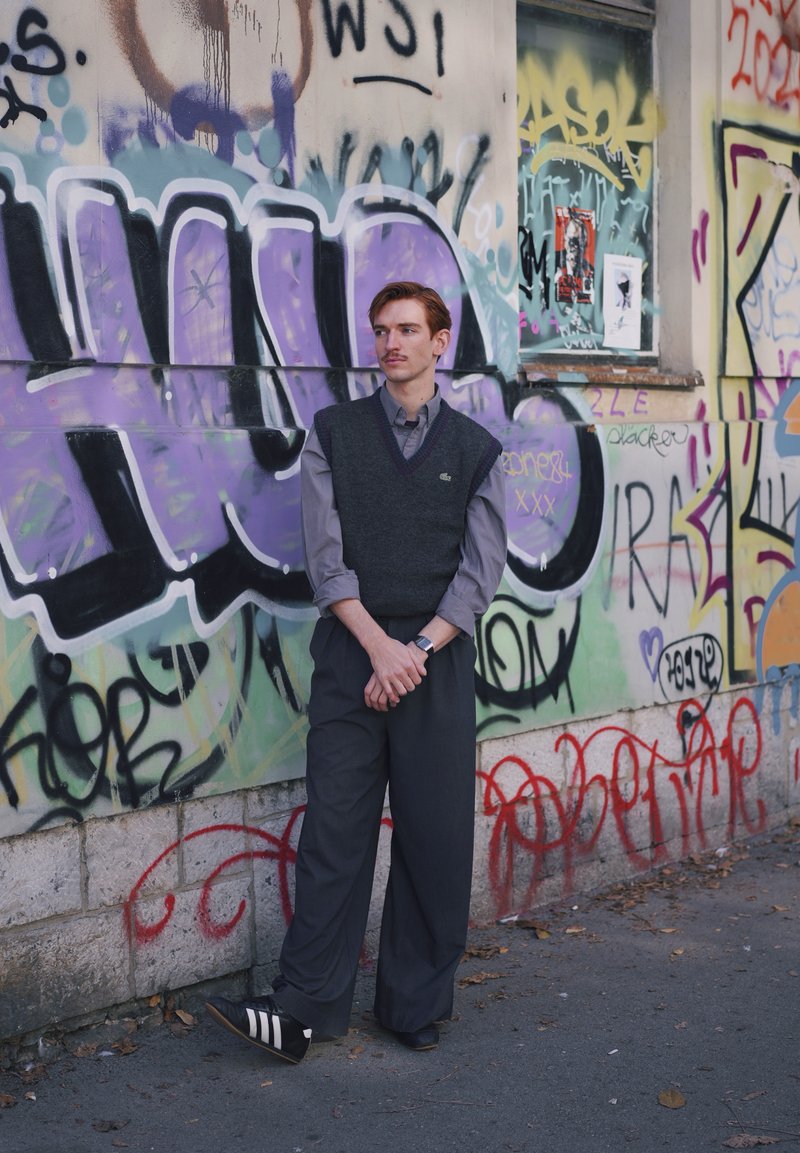 Homem jovem com camisa cinza, colete escuro e calças largas, encostado numa parede coberta de grafite numa rua urbana.