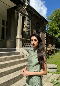Jasnozielona sukienka z drapowaniem, o wysokim dekolcie i miękkiej teksturze. Modelka stoi na kamiennych schodach, w tle widać ceglaną budowlę i zieleń.