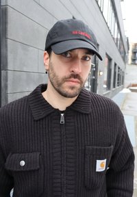 Άνδρας με γένια, φορώντας μαύρο καπέλο και σκούρο πλεκτό πουλόβερ με φερμουάρ, στέκεται έξω από αστικό κτίριο σε συννεφιασμένο καιρό.