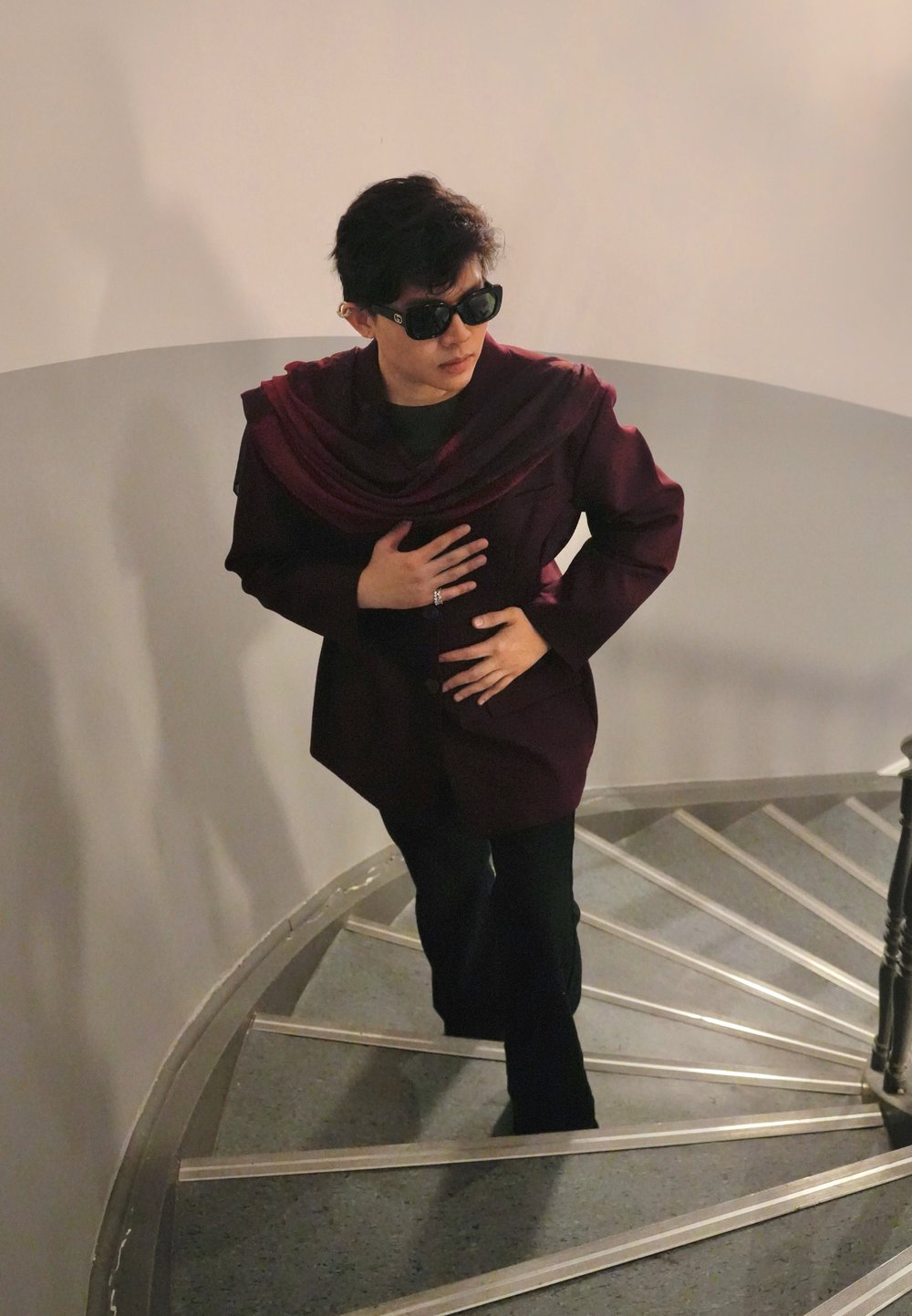 Personne portant un blazer bordeaux foncé avec un col drapé, un haut noir et un pantalon noir évasé, descendant un escalier en spirale.