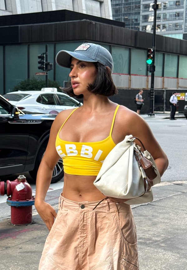Gul crop top med hvite bokstaver, kombinert med beige vide shorts. Holder en hvit veske, iført en lyseblå baseball cap.