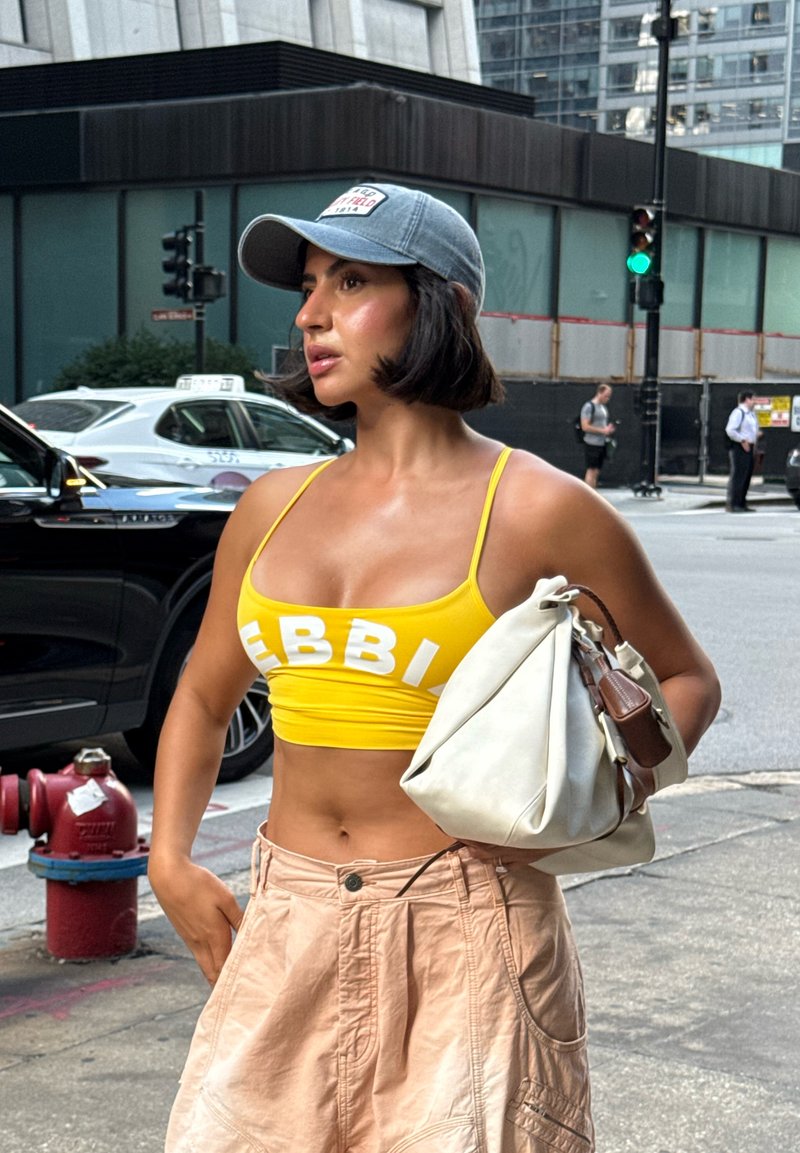 Gelbes Crop-Top mit weißer Schrift, kombiniert mit beigen weit geschnittenen Shorts. Hält eine weiße Tasche und trägt eine hellblaue Baseballmütze.