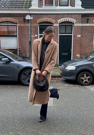 Abrigo largo beige sobre una camisa negra, jeans anchos oscuros y botines puntiagudos. Sostiene un bolso tejido negro con textura. Fondo urbano.