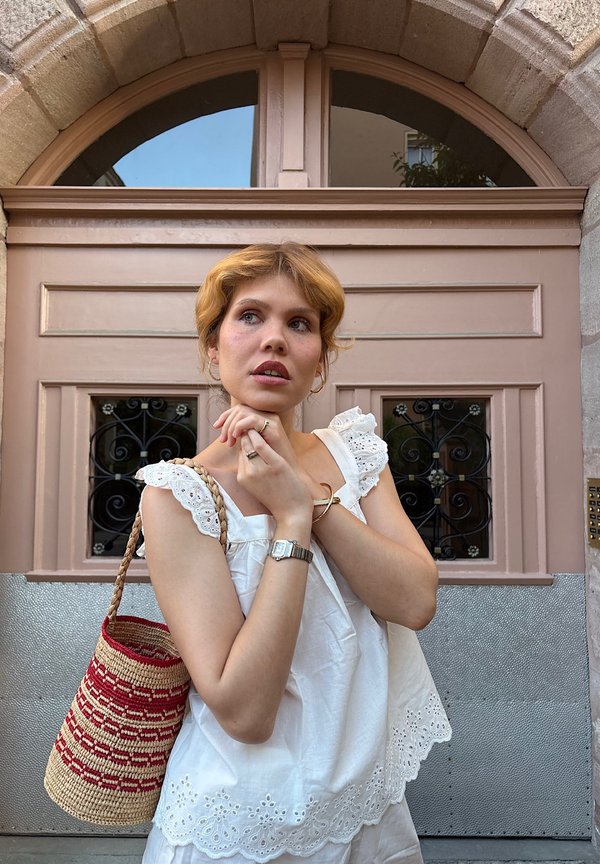 Jeune femme aux cheveux blonds courts portant un haut blanc en broderie anglaise et tenant un sac tissé rouge et beige, debout devant une grande porte rose.
