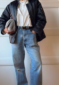 Μαύρο επενδυμένο μπουφάν bomber πάνω από λευκό πουκάμισο, συνδυασμένο με ανοιχτό μπλε τζιν και γκρίζο υφή αξεσουάρ στο χέρι.