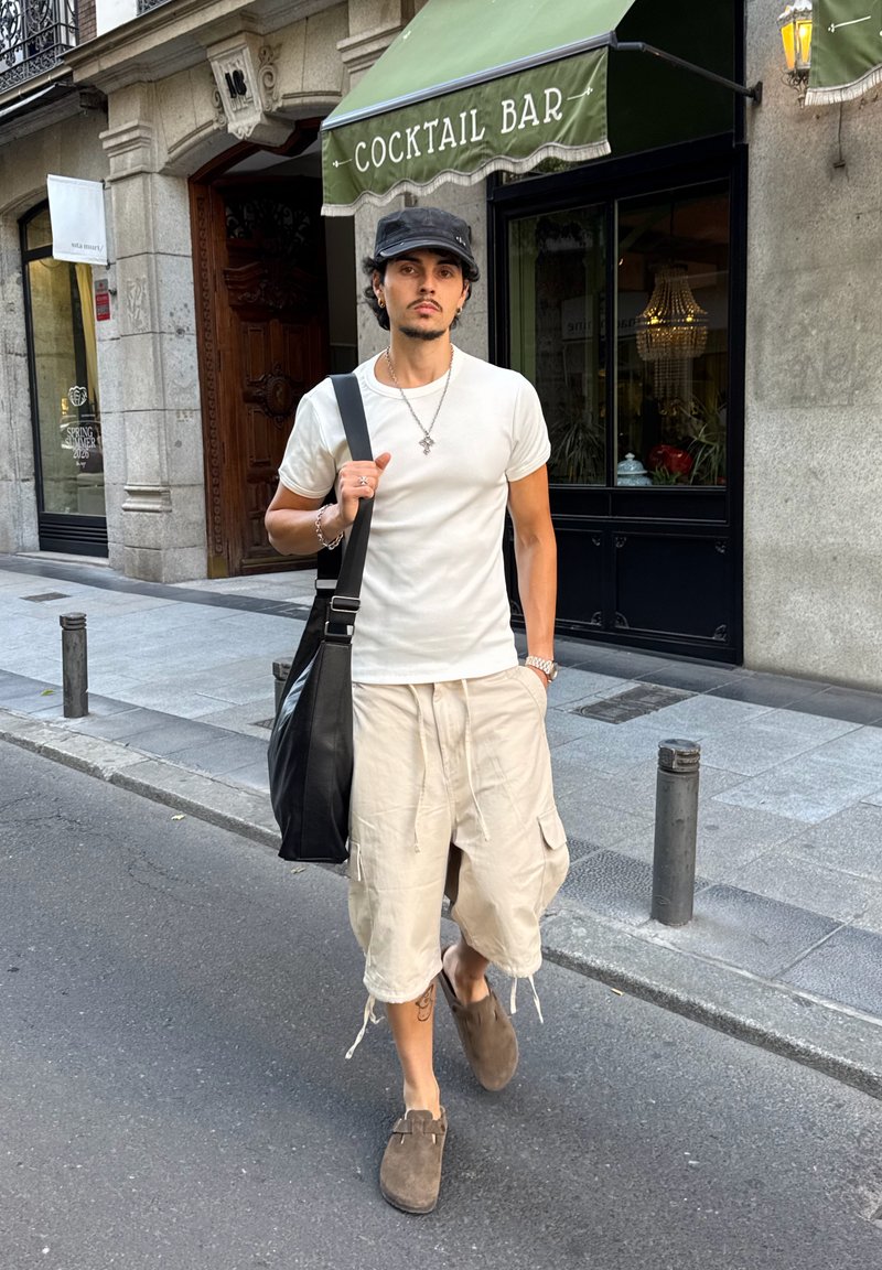 Jeune homme portant un t-shirt blanc, un short cargo beige, des chaussures marron et une casquette noire, marchant dans une rue de la ville près d'un bar à cocktails.