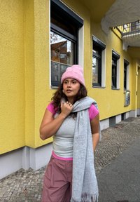 Écharpe grise drapée sur un haut rose et gris, assortie à un bonnet en laine rose. En arrière-plan, un mur jaune et des fenêtres rectangulaires.