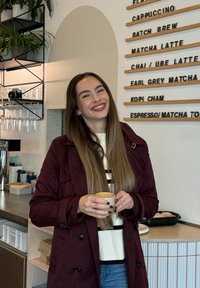 Smilende kvinne med langt hår iført en bordeauxfarget frakk holder en kopp kaffe inne i en kafé med en meny på veggen bak henne.