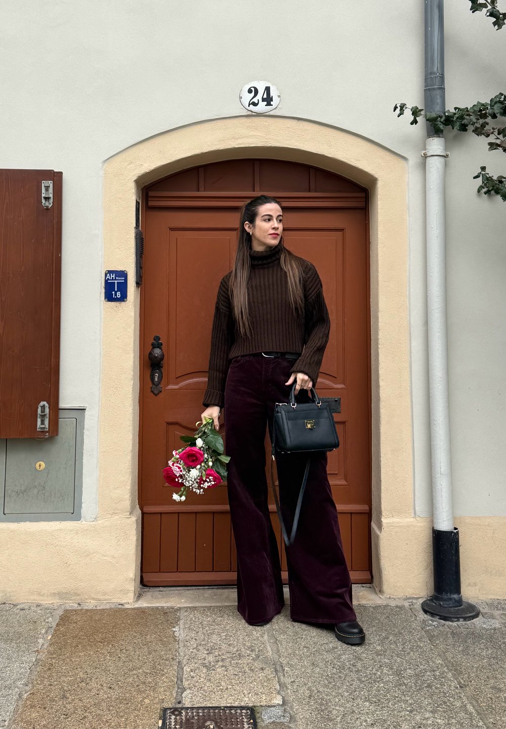 Brown ribbed turtleneck jumper, burgundy wide-leg corduroy trousers, black handbag, holding a bouquet of red roses and gypsophila, standing by a door.
