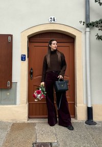 Brown ribbed turtleneck jumper, burgundy wide-leg corduroy trousers, black handbag, holding a bouquet of red roses and gypsophila, standing by a door.