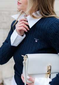 Pulover bleumarin cu model tricotat cu torsade, cu un guler alb, purtând un mic logo GANT. Țin o geantă albă cu detalii aurii.