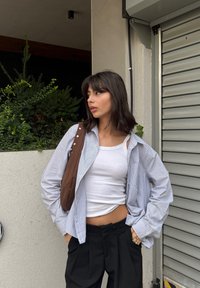 Camisa riscada azul claro e branca, usada aberta sobre uma t-shirt branca. Mala de ombro castanha com formato arredondado e botões. Calças pretas.