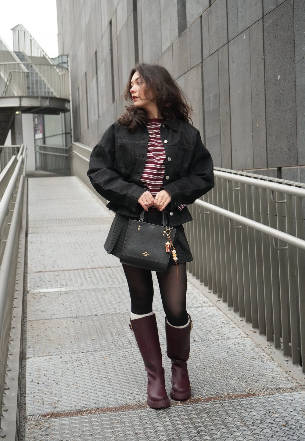Jeune femme portant une veste noire, une chemise rayée, une jupe plissée, des collants noirs et des bottes bordeaux, tenant un sac à main noir sur une passerelle métallique.