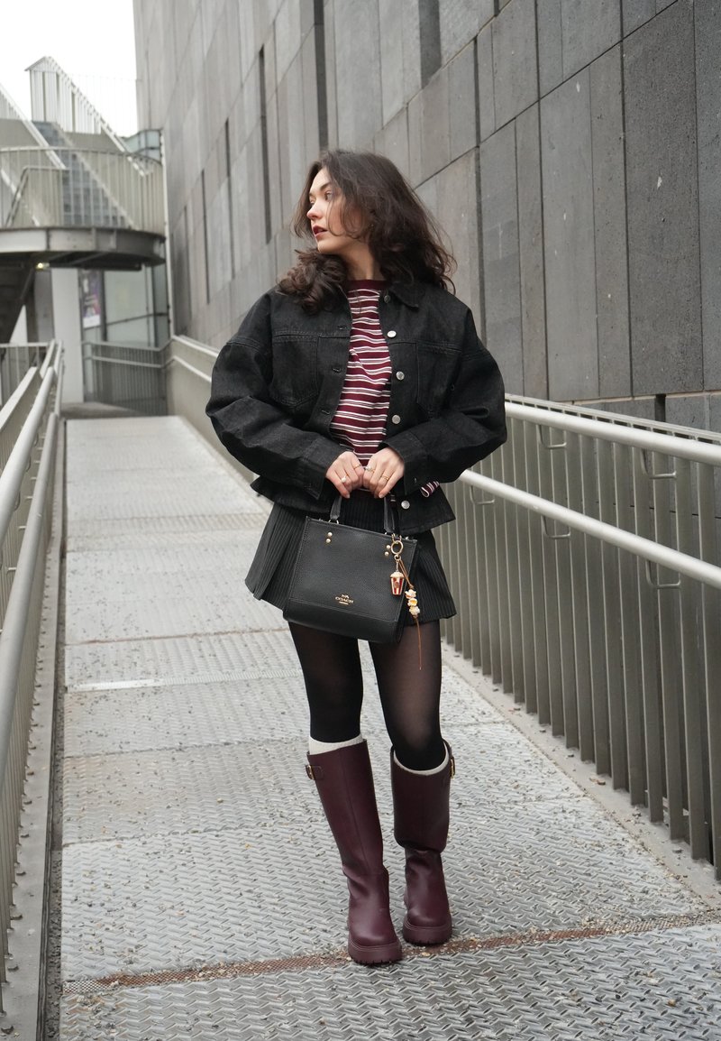 Giovane donna con giacca nera, camicia a righe, gonna plissettata, calze nere e stivali color bordeaux che tiene una borsa nera su una passerella metallica.