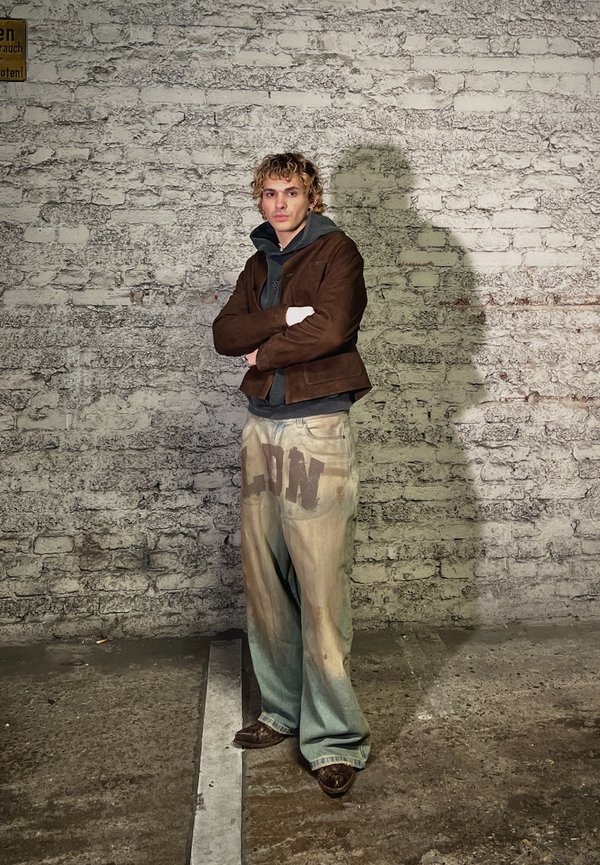 Νεαρό άτομο με ξανθά κατσαρά μαλλιά στέκεται με τα χέρια σταυρωμένα, φορώντας καφέ μπουφάν, μαύρη κουκούλα και φαρδιά μπεζ παντελόνια με σκούρα γράμματα.