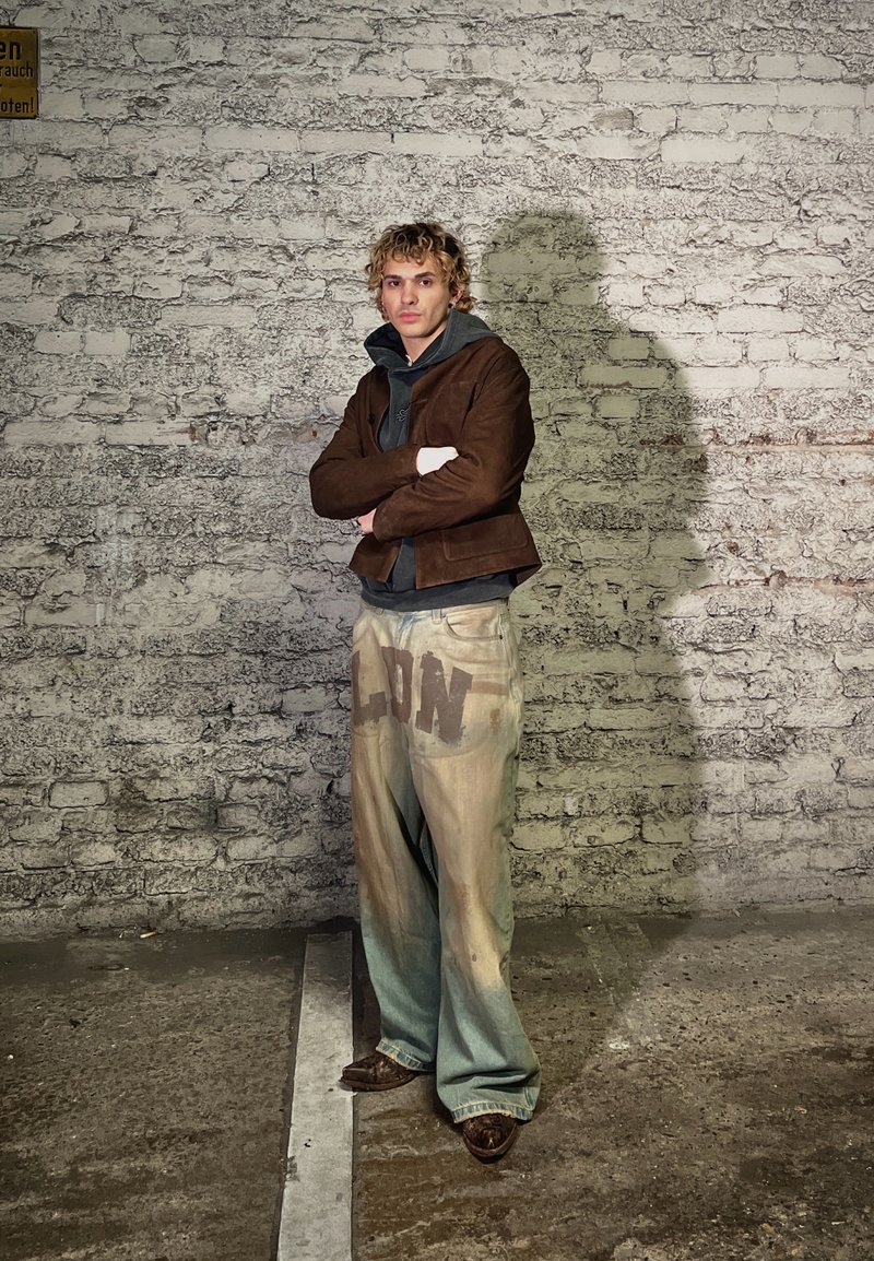 Jeune personne aux cheveux blonds bouclés, debout les bras croisés, portant une veste marron, un sweat à capuche noir et un pantalon large beige avec des lettres foncées.