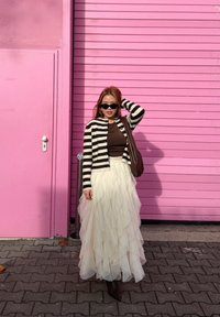 Cardigan rayé noir et blanc, haut marron et jupe en tulle ivoire superposée. Bottes foncées et lunettes de soleil complètent la tenue sur un fond rose.