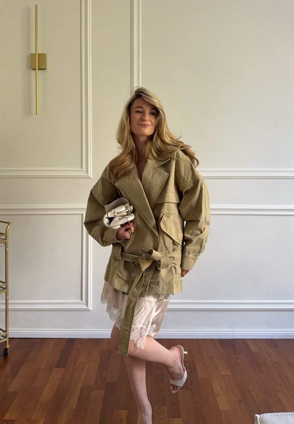 Žena v nadměrné khaki bundě a šatech s krajkovým lemem drží malou bílou kabelku a stojí na jedné noze v minimalistické místnosti s dřevěnou podlahou.