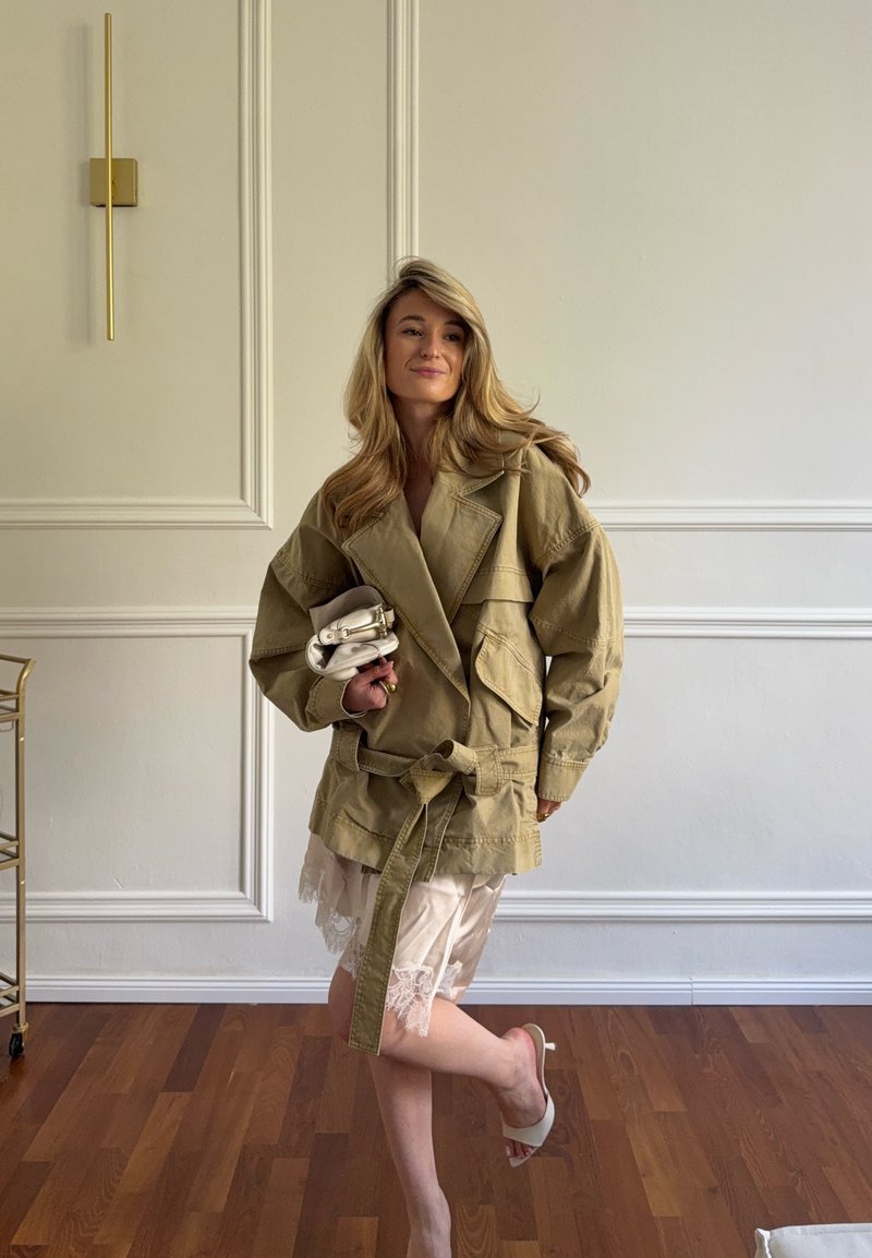 Femme en veste kaki oversize et robe bordée de dentelle tenant un petit sac blanc, debout sur une jambe dans une pièce minimaliste avec un sol en bois.