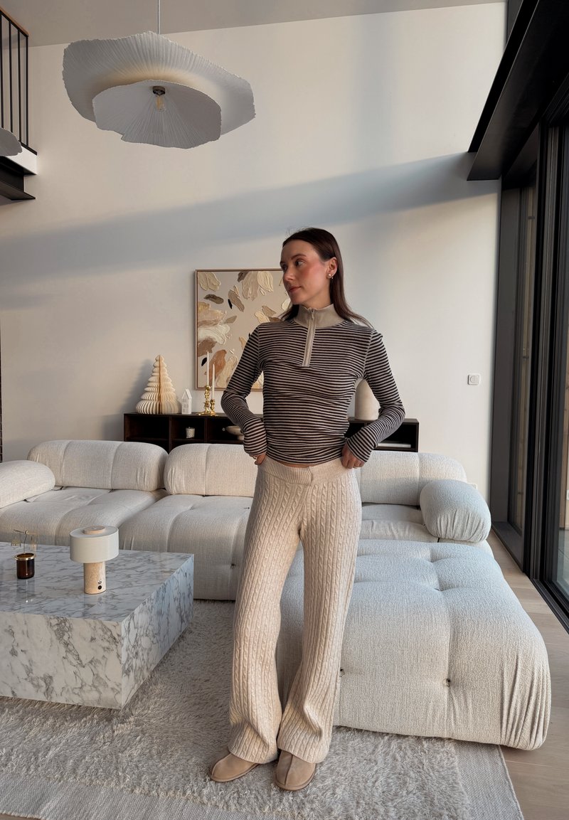 Striped long-sleeve top with a zip collar, light beige cable-knit trousers, and beige slip-on shoes, set in a modern interior with a marble table.