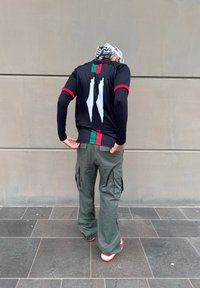 Μαύρη μπλούζα με μακρύ μανίκι και κόκκινες λεπτομέρειες, με ένα λευκό σχέδιο στην πλάτη; πράσινα cargo παντελόνια; κόκκινα και λευκά αθλητικά παπούτσια.