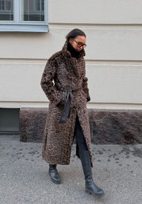 Manteau imprimé léopard avec taille ceinturée, texture en fausse fourrure, col haut et fentes latérales, associé à des bottes en cuir noir montant jusqu'aux genoux.