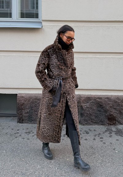 Abrigo de estampado de leopardo con cintura ceñida, textura de piel sintética, cuello alto y aberturas laterales, combinado con botas de cuero negras hasta la rodilla.