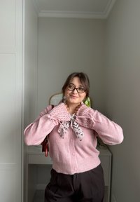 Femme portant des lunettes et un pull rose en tricot avec des poignets en dentelle, ajustant une écharpe dans une pièce minimaliste aux couleurs claires.