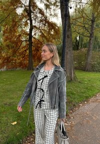 Pelēka ādas motociklistu jaka pārbaudītai blūzei un atbilstošiem biksēm. Apģērbs ir papildināts ar gaišas krāsas somiņu.