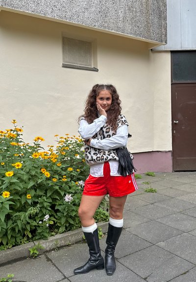 Chaleco de estampado de leopardo sobre camisa blanca, pantalones cortos de satén rojos, botas altas negras con acentos blancos, de pie frente a flores y un edificio.