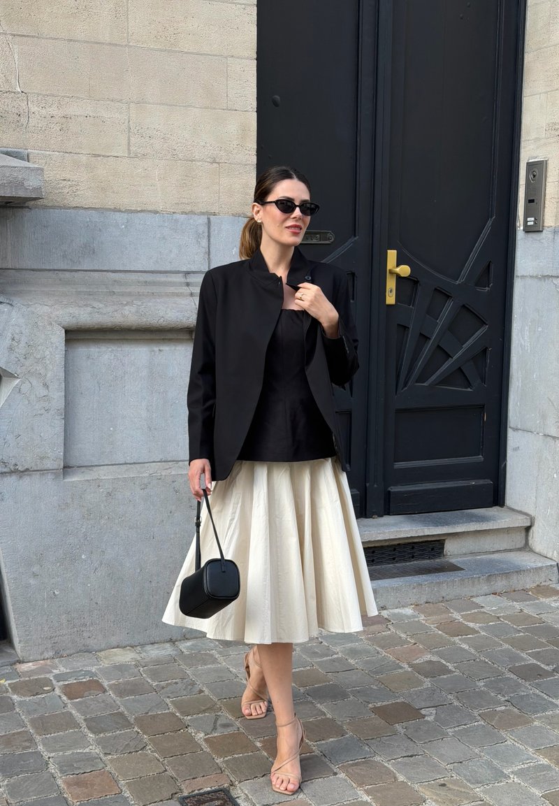 Woman wearing black jacket, black top, cream pleated skirt, and sandals holding a black handbag standing on cobblestone street by a dark door.