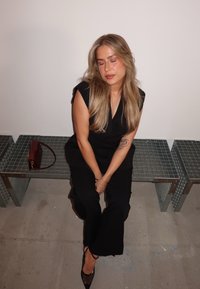 Svart, ärmlös jumpsuit med v-ringad halsringning och vida ben. Långt, vågigt blont hår. Lätt smink, sittande med en liten vinröd handväska i närheten.