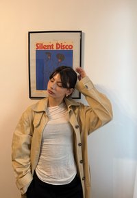 Beżowy płaszcz przeciwdeszczowy z dużym kołnierzem i guzikami na białej koszuli, zestawiony z czarnymi spodniami. W tle oprawiony plakat z napisem "Silent Disco".