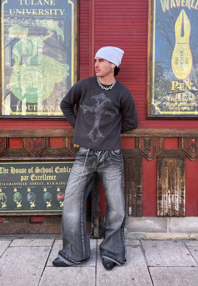 Suéter negro con un diseño de cruz gris, jeans de pierna ancha grises desgastados con detalle de cadena, botas negras y un gorro blanco. El fondo presenta carteles antiguos.
