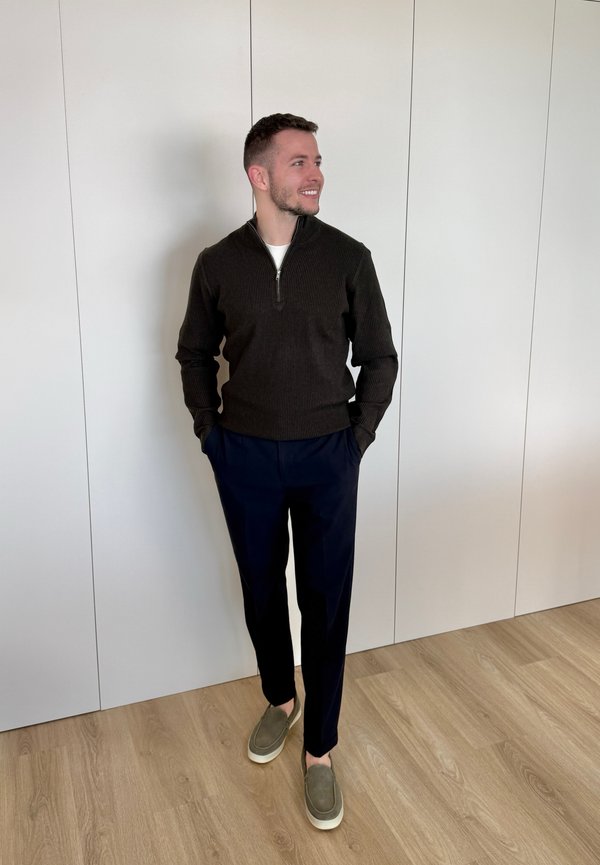 Mann steht drinnen an einer weissen Wand, trägt einen dunklen Reissverschluss-Pullover, marineblaue Hosen und grüne Slipper, Hände in den Taschen, lächelnd und schaut nach rechts.