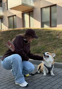 Jonge vrouw in een pet hurkt op de stoep, glimlachend en een blije Corgi aaien die een harnas draagt, buiten naast een grashelling en gebouwen.